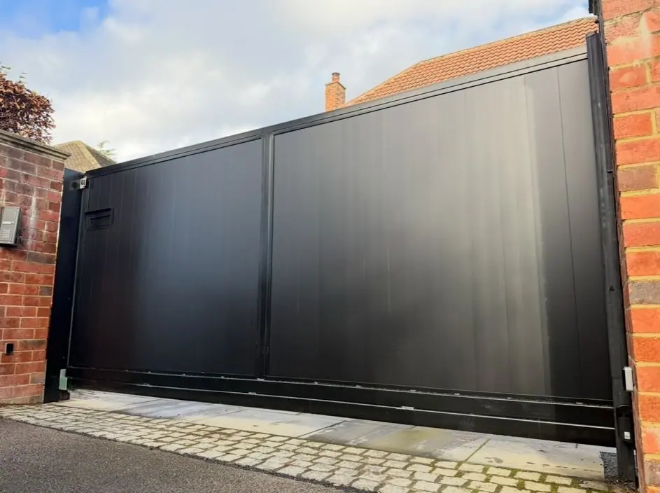Black boarded electric sliding gate closed across a setted driveway, against a red brick wall