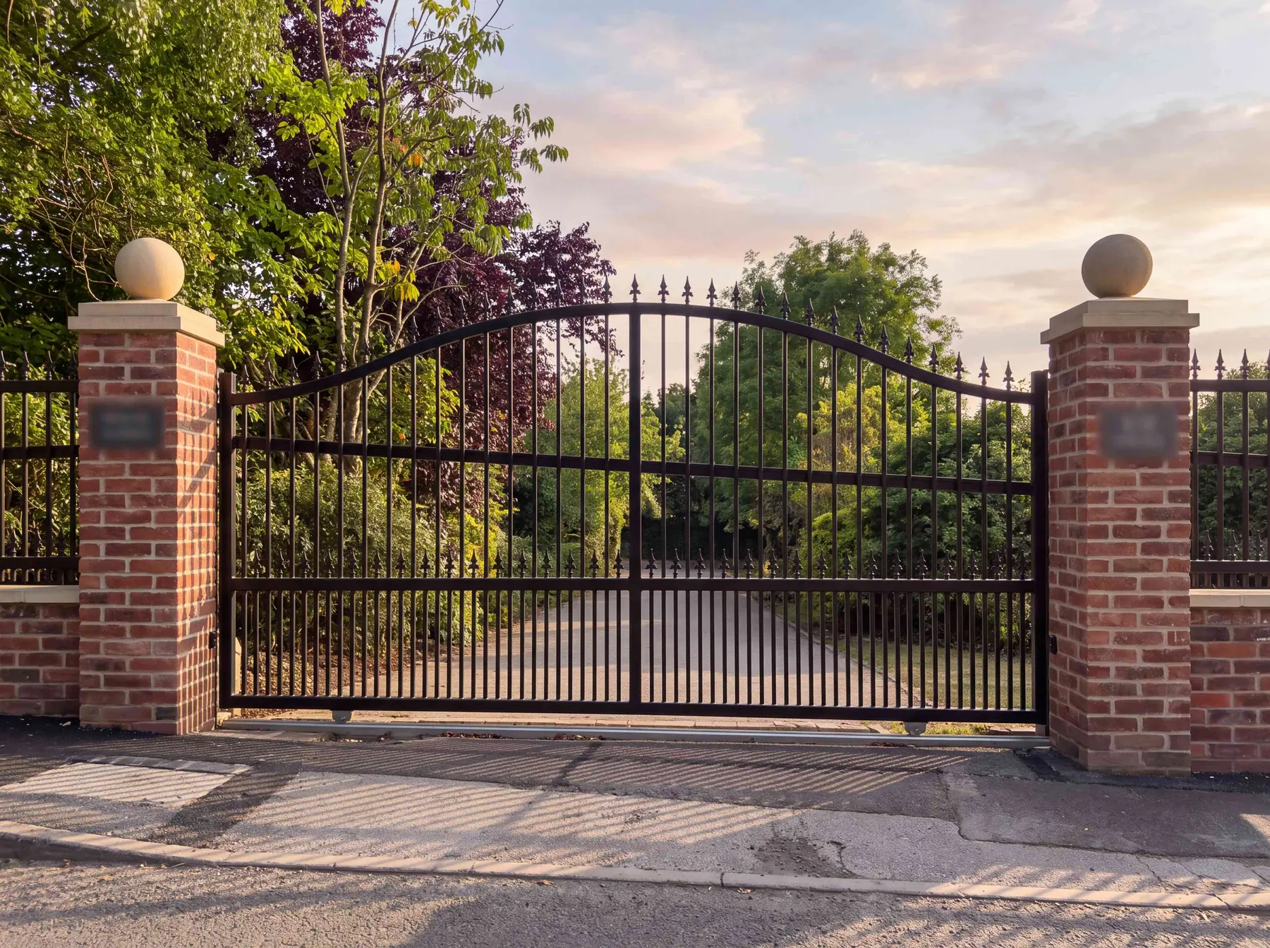 Arched wrought iron electric gates between red brick pillars with ball finials