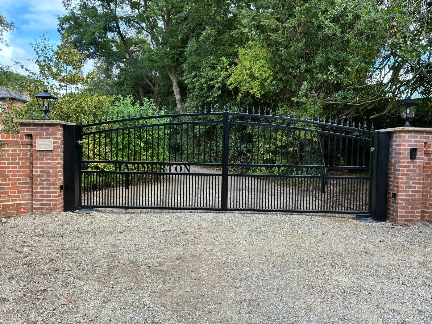 Arched wrought iron electric gates with the property name worked into the ironwork, between brick piers with coach lanterns