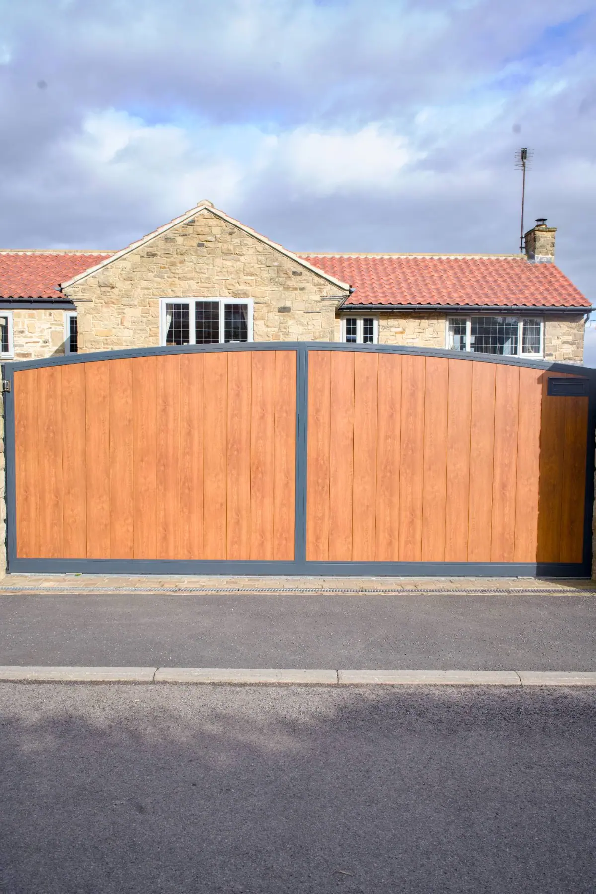 Curved-top electric driveway gates with oak-effect infill and a dark grey frame, at a Yorkshire stone house