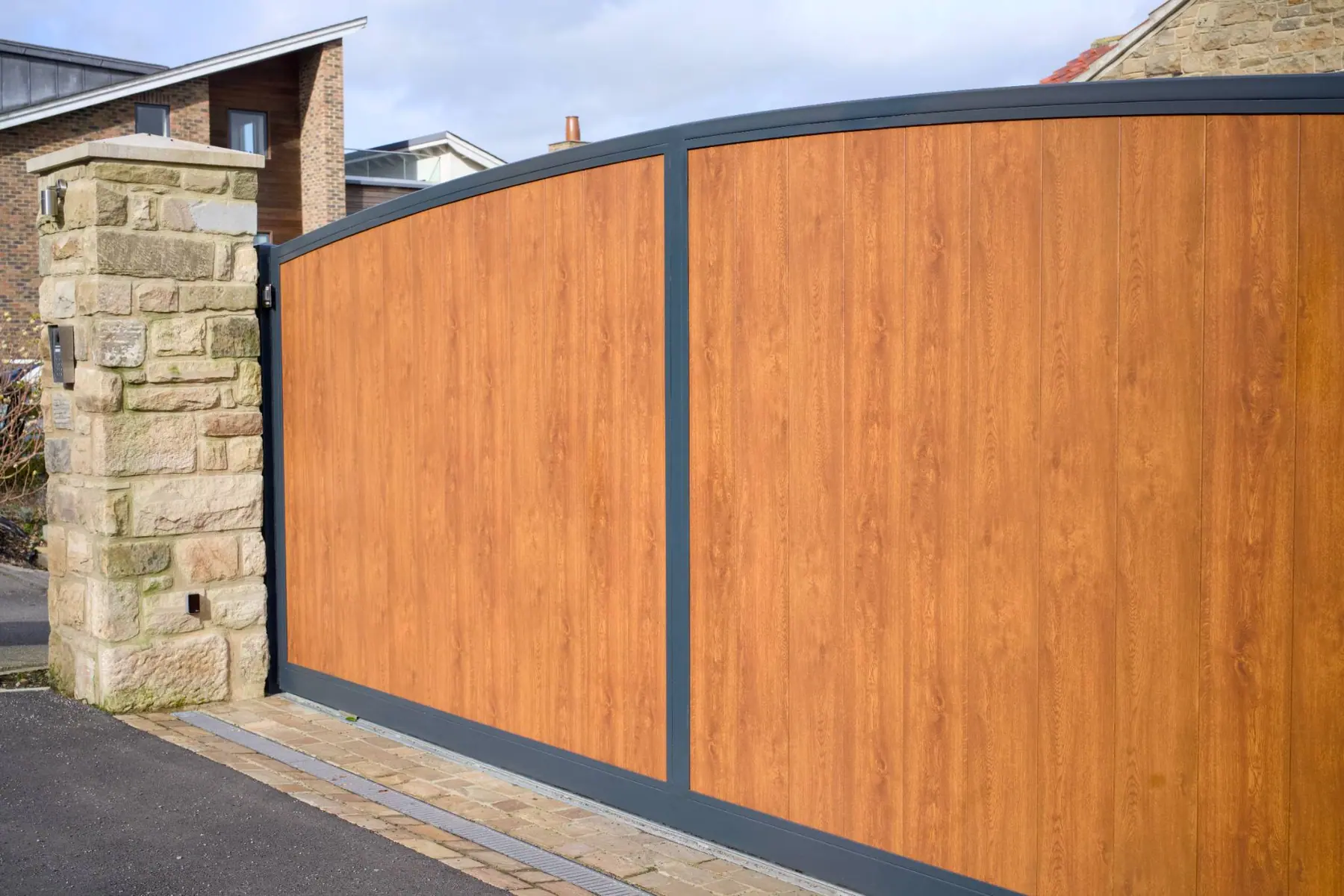 Curved-top electric driveway gates with oak-effect infill and a dark grey frame, at a Yorkshire stone house