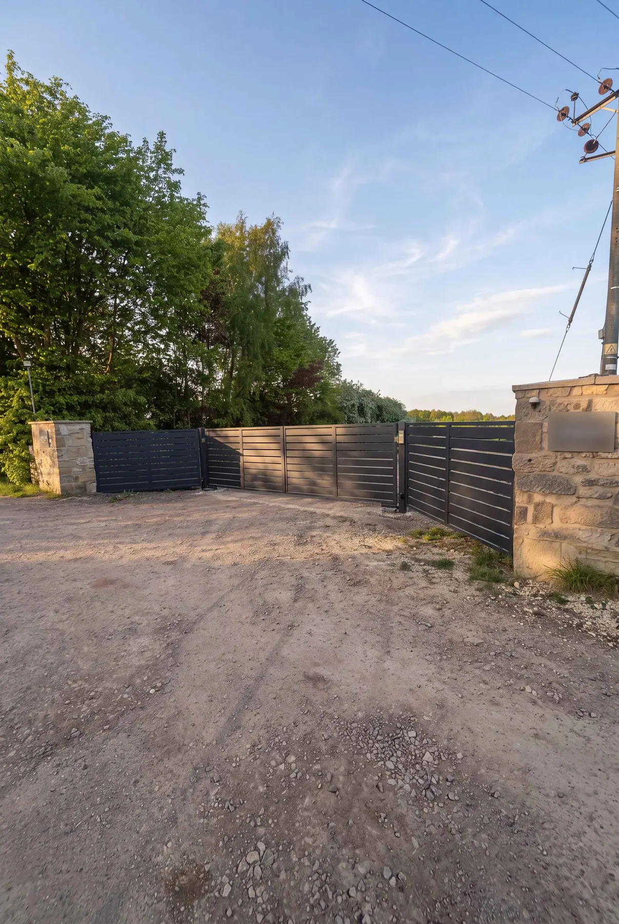 Dark slatted electric gate on a gravel driveway between stone pillars