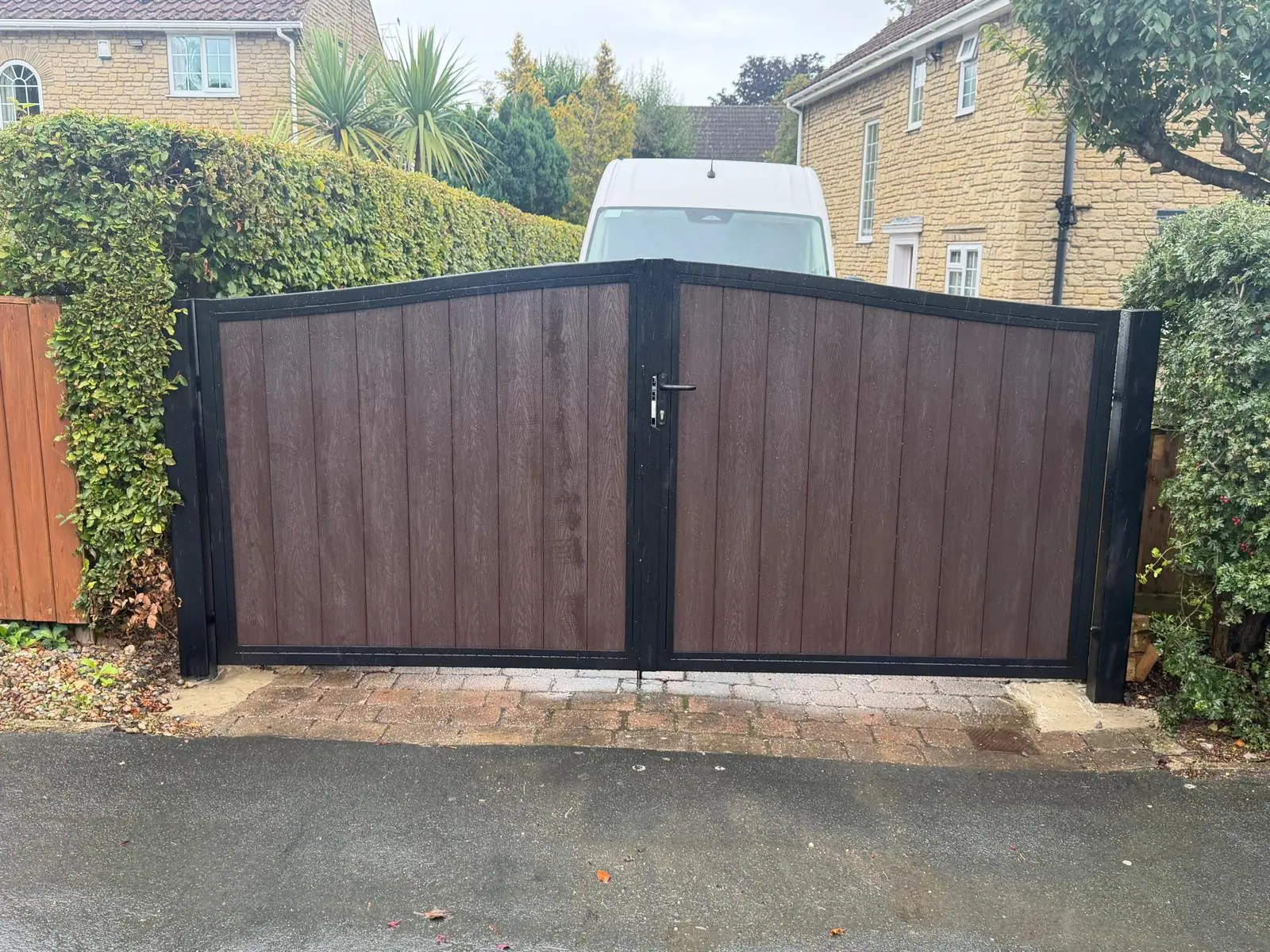 Wood-effect composite manual gates with a black frame and a lever handle, between a hedge and a fence